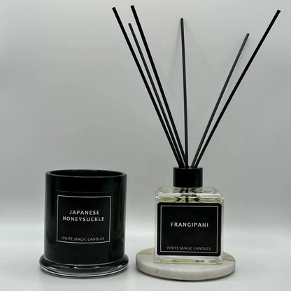 Two black glass jars with white labels reading "JAPANESE HONEYSUCKLE" and "FRANGIPANI" sit on a white surface. The left jar is a wax candle with a black lid. The right jar is a reed diffuser with black reeds.