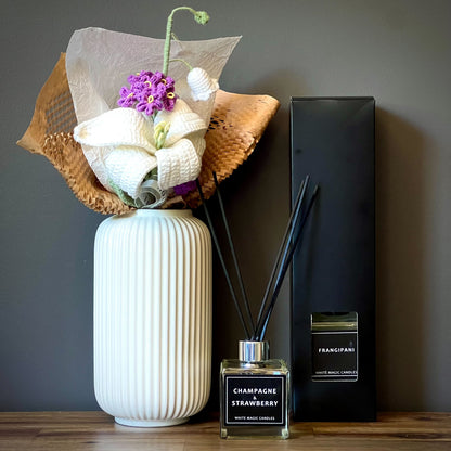 A white vase with a floral arrangement and a black reed diffuser candles bottle with a white label reading "FRANGIPANI" sits on a wooden surface.