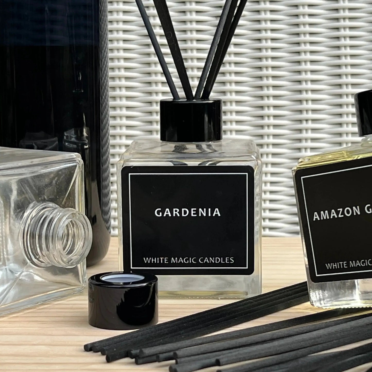 A collection of glass Buy reed diffuser bottles with black labels and black caps, surrounded by reeds. The labels read "GARDENIA," "AMAZON GRAPE