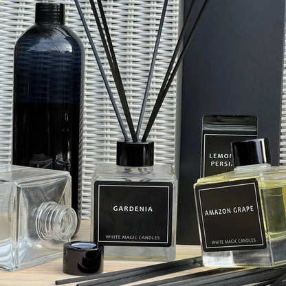 A collection of glass reed diffuser bottles with black labels and black caps, surrounded by reeds. The labels read "GARDENIA," "AMAZON GRAPE," and "LEMON PERSIA."