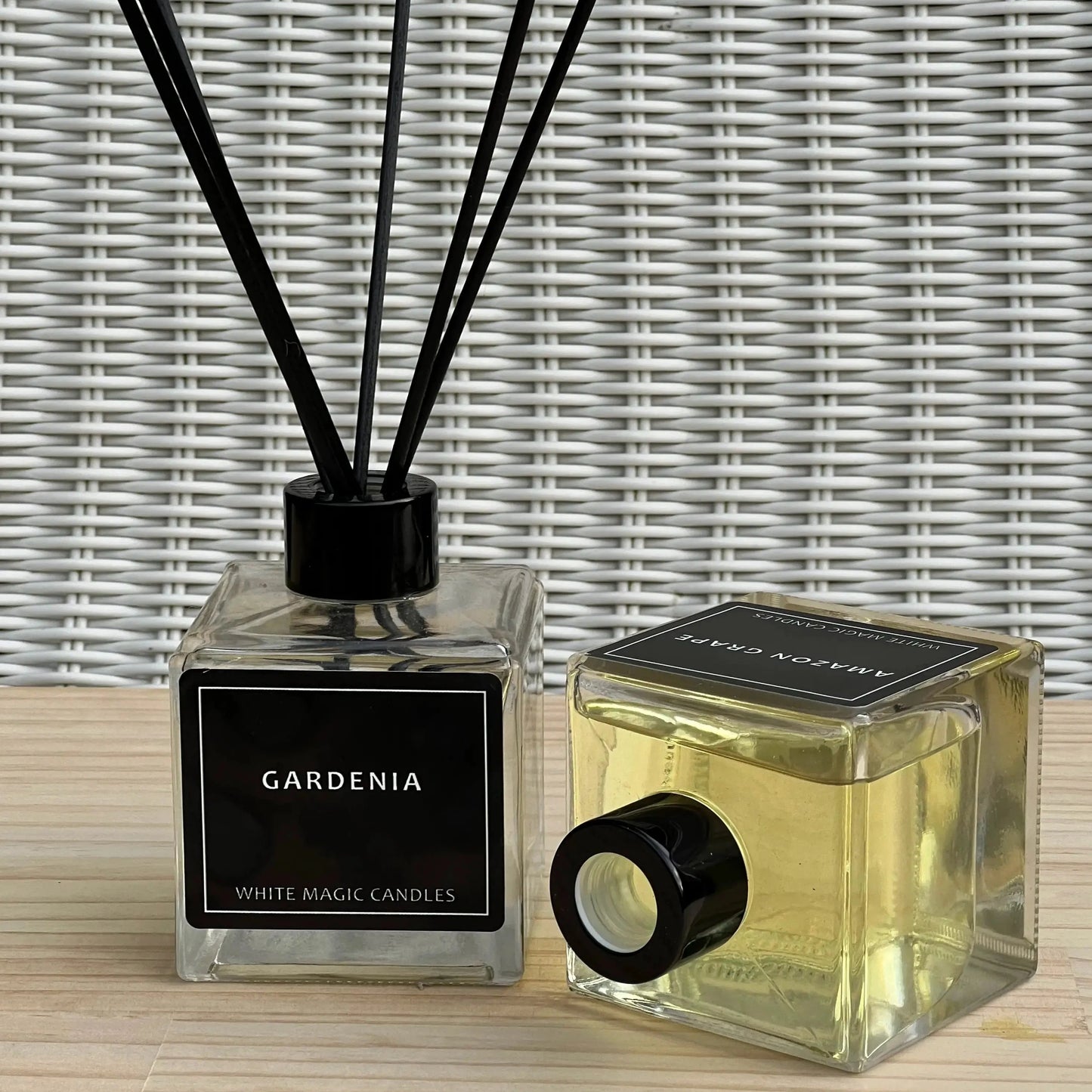 Two black glass jars with white labels reading gardenia  The left jar is a wax candle with a black lid. The right jar is a reed diffuser with black reeds.