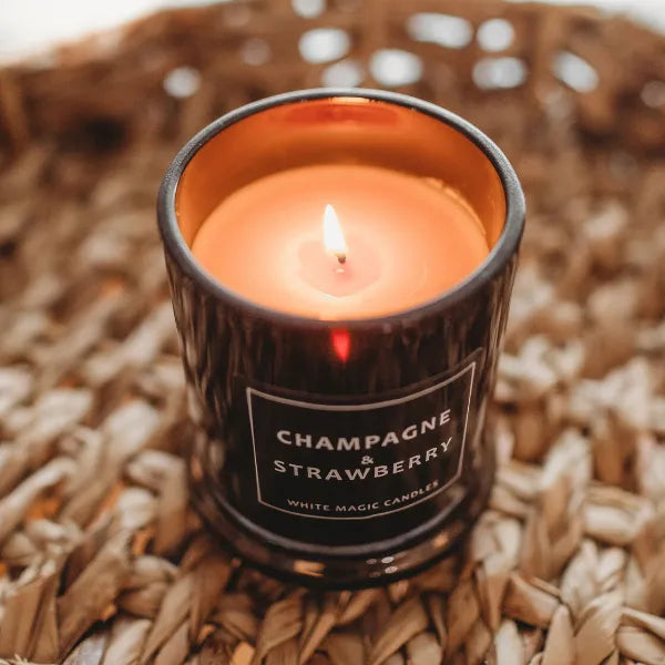 Black glass candle jar with a white label reading "Champagne & Strawberry" and "White Magic Candles." The buy soy wax candle is lit and sitting on a woven surface.