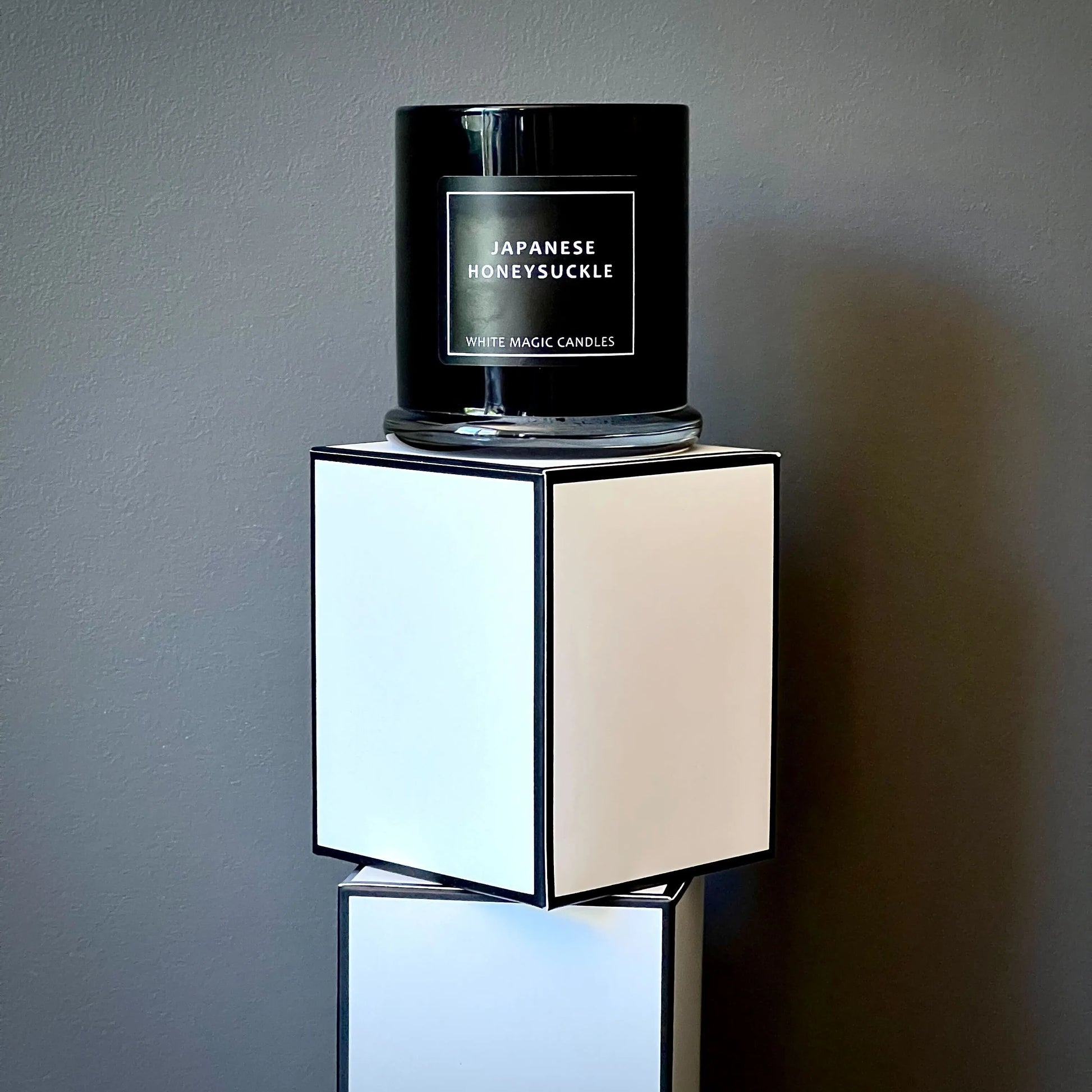 Black glass candle jar with a white label reading "JAPANESE HONEYSUCKLE" and "WHITE MAGIC CANDLES" sitting on top of two stacked white boxes.