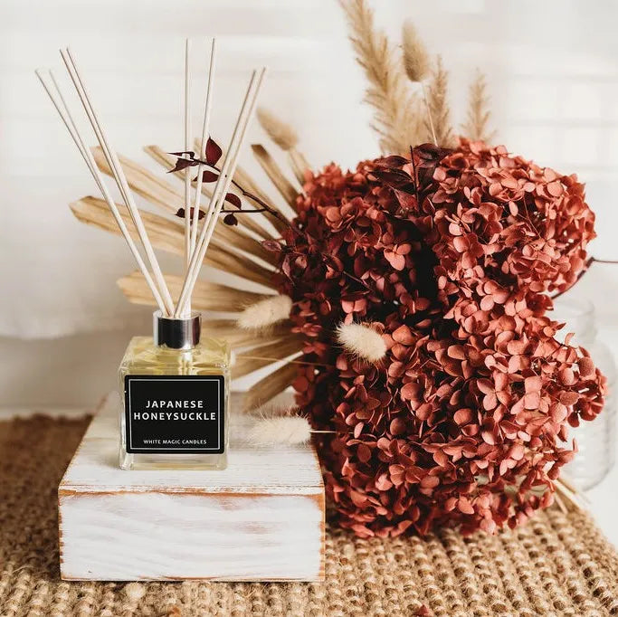 "A square glass reed diffuser bottle with a black label reading 'JAPANESE HONEYSUCKLE' sits on a white wooden box, surrounded by dried flowers. Buy reed diffusers."